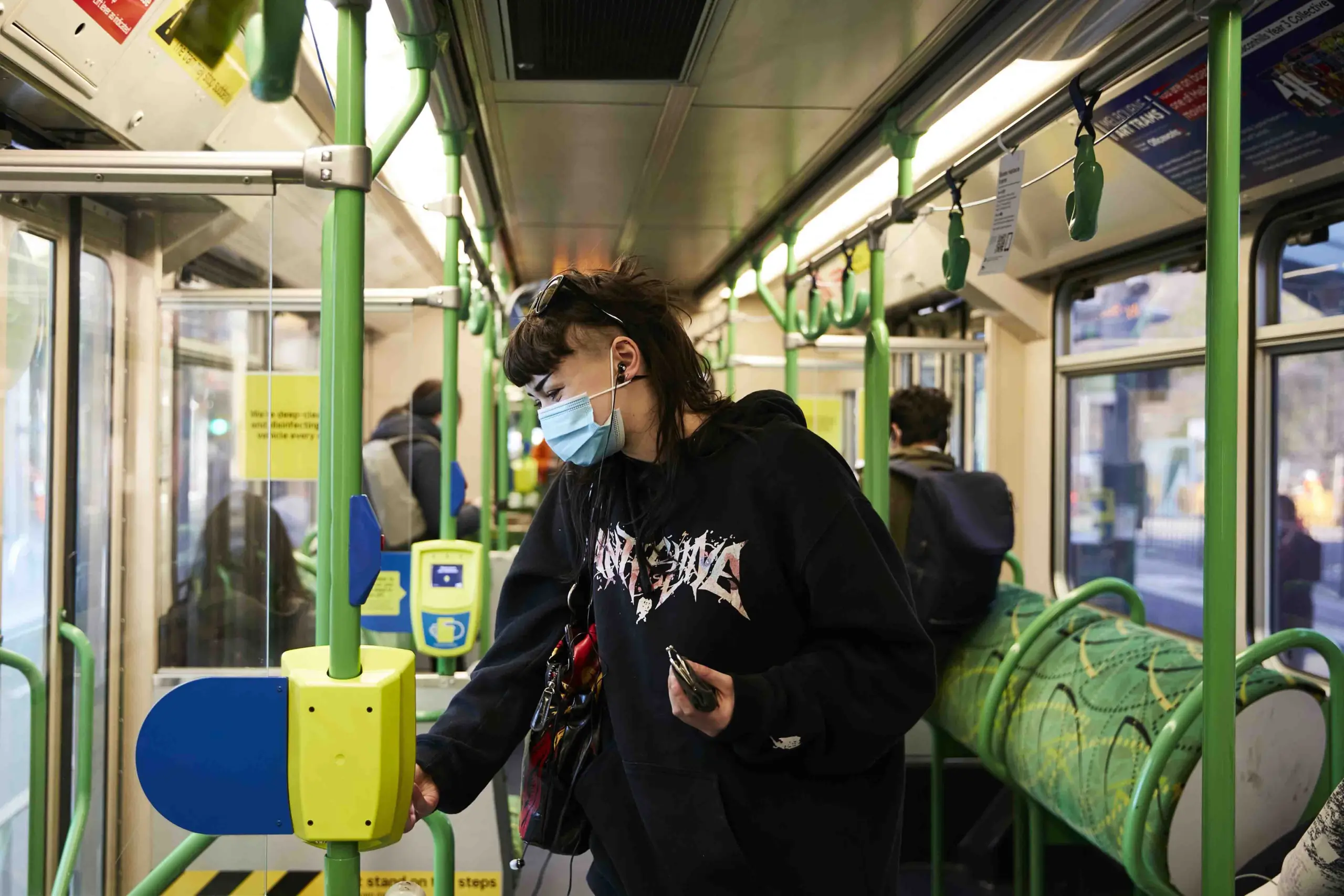 A young woman on a tram tapping her myki on the reader. She is wearing a facemask.