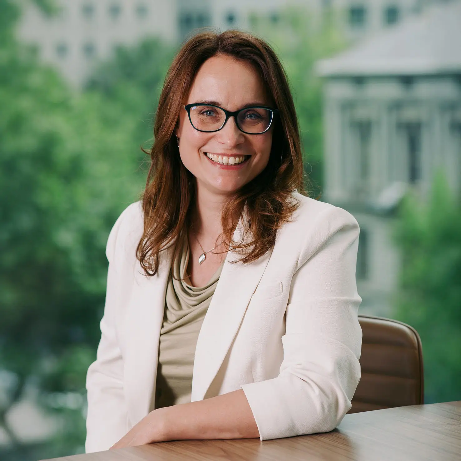 Professional portrait of smiling woman seated at desk, with blurred trees and buildings in the background.