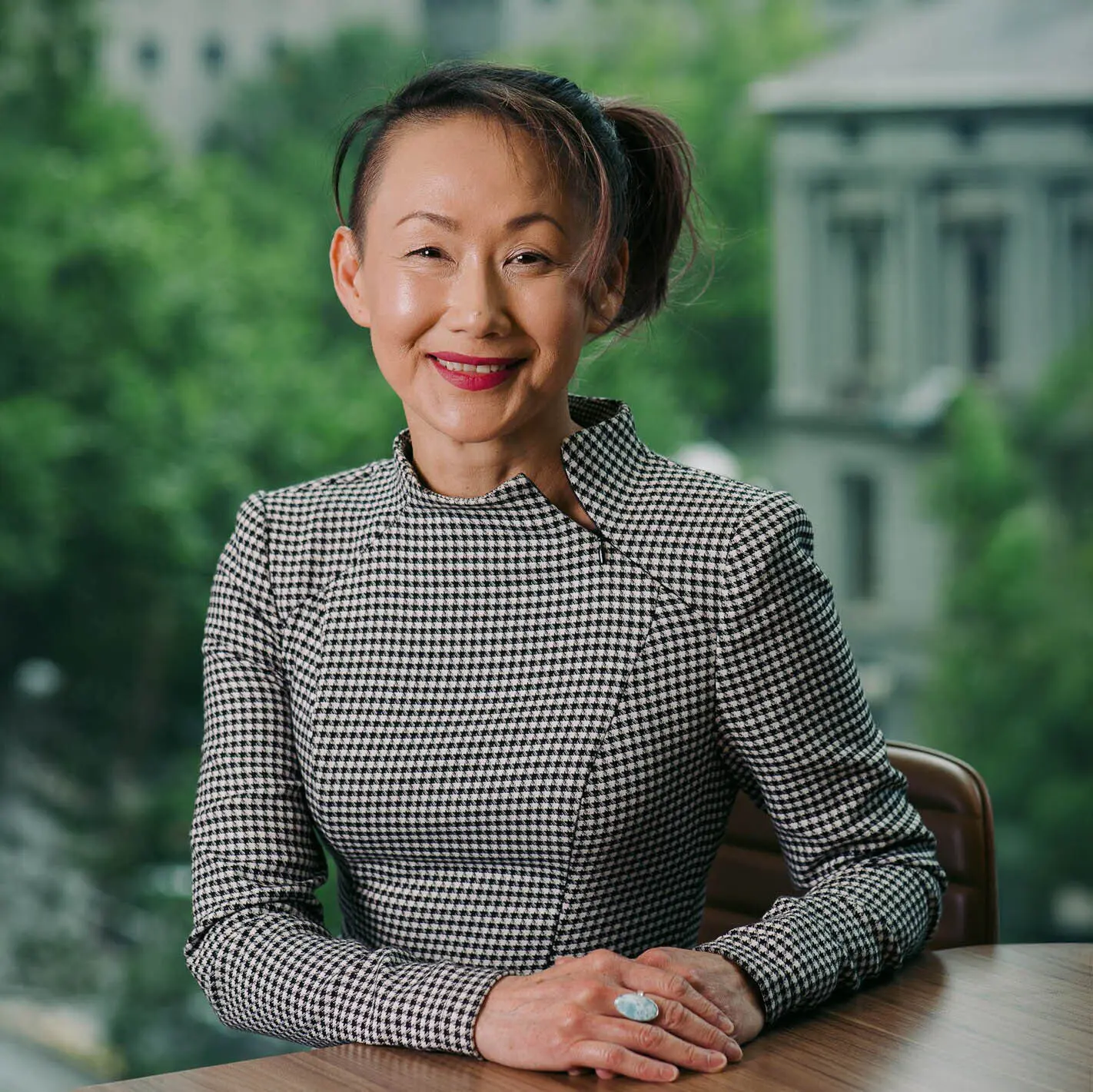 Professional portrait of smiling woman seated at desk, with blurred trees and buildings in the background.