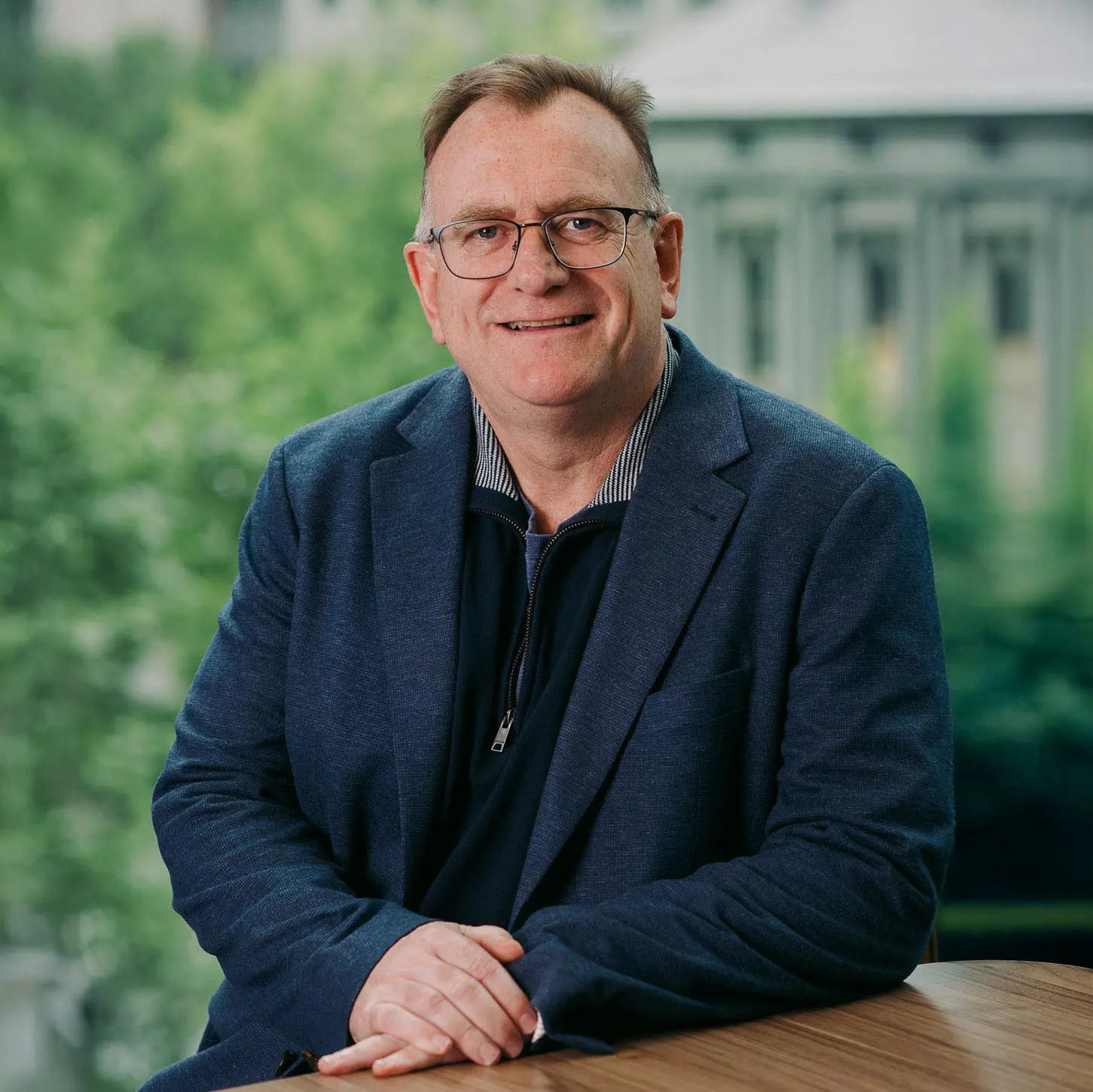 Professional portrait of smiling man seated at desk, with blurred trees and building in the background.