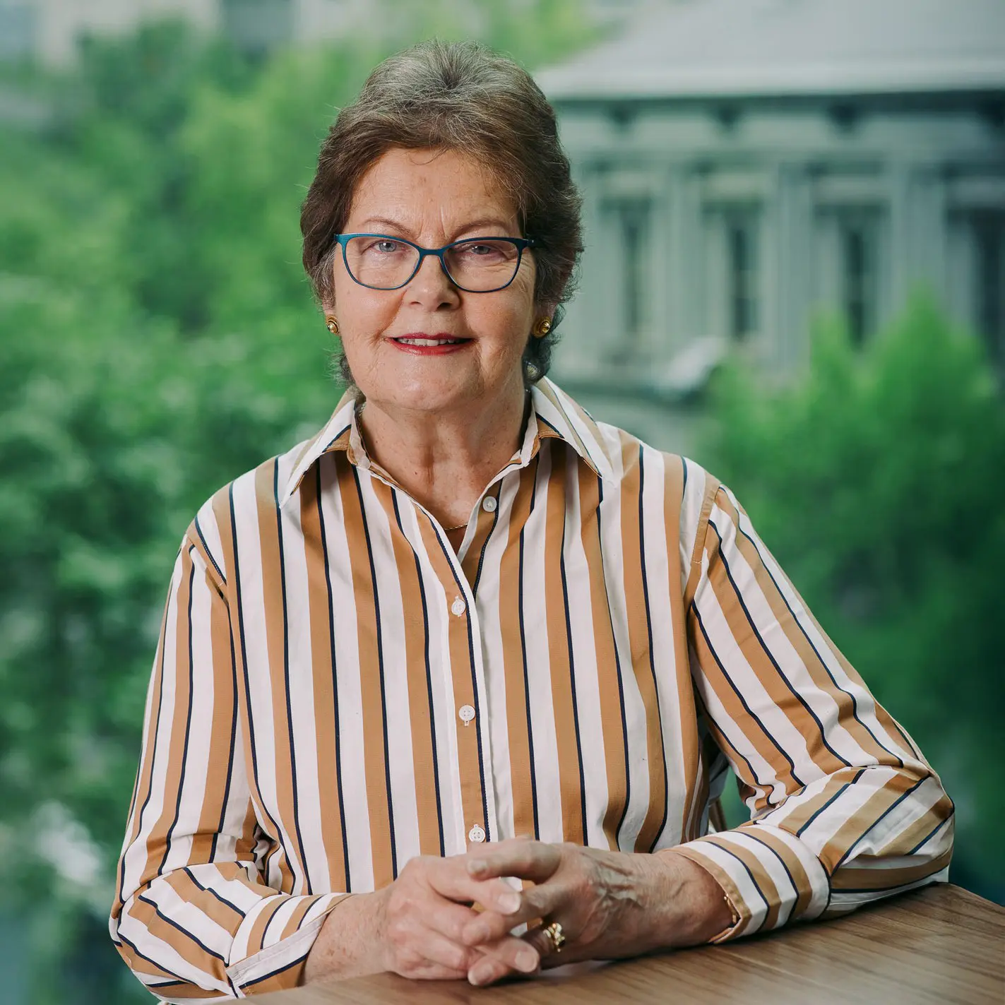 Professional portrait of smiling woman seated at desk, with blurred trees in the background.
