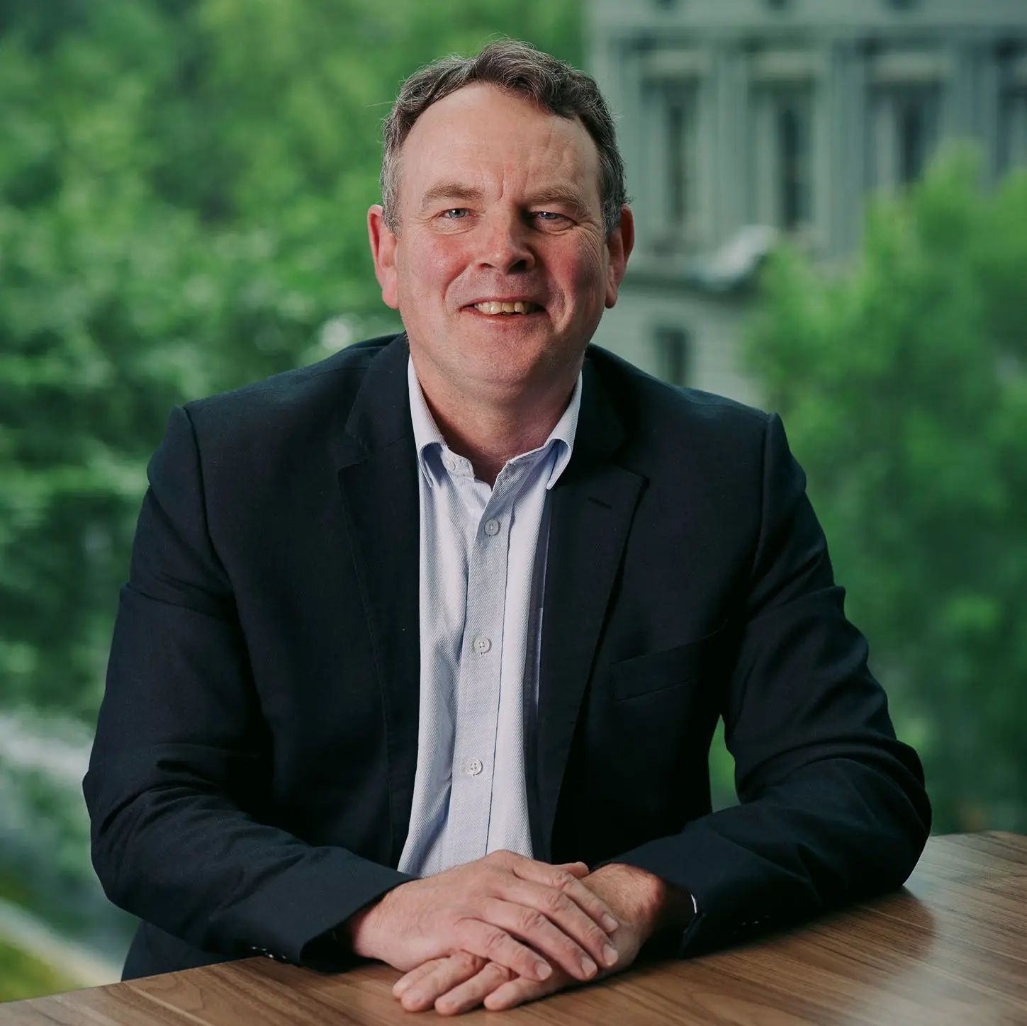 Professional portrait of smiling man seated at desk, with blurred trees and garden in the background.