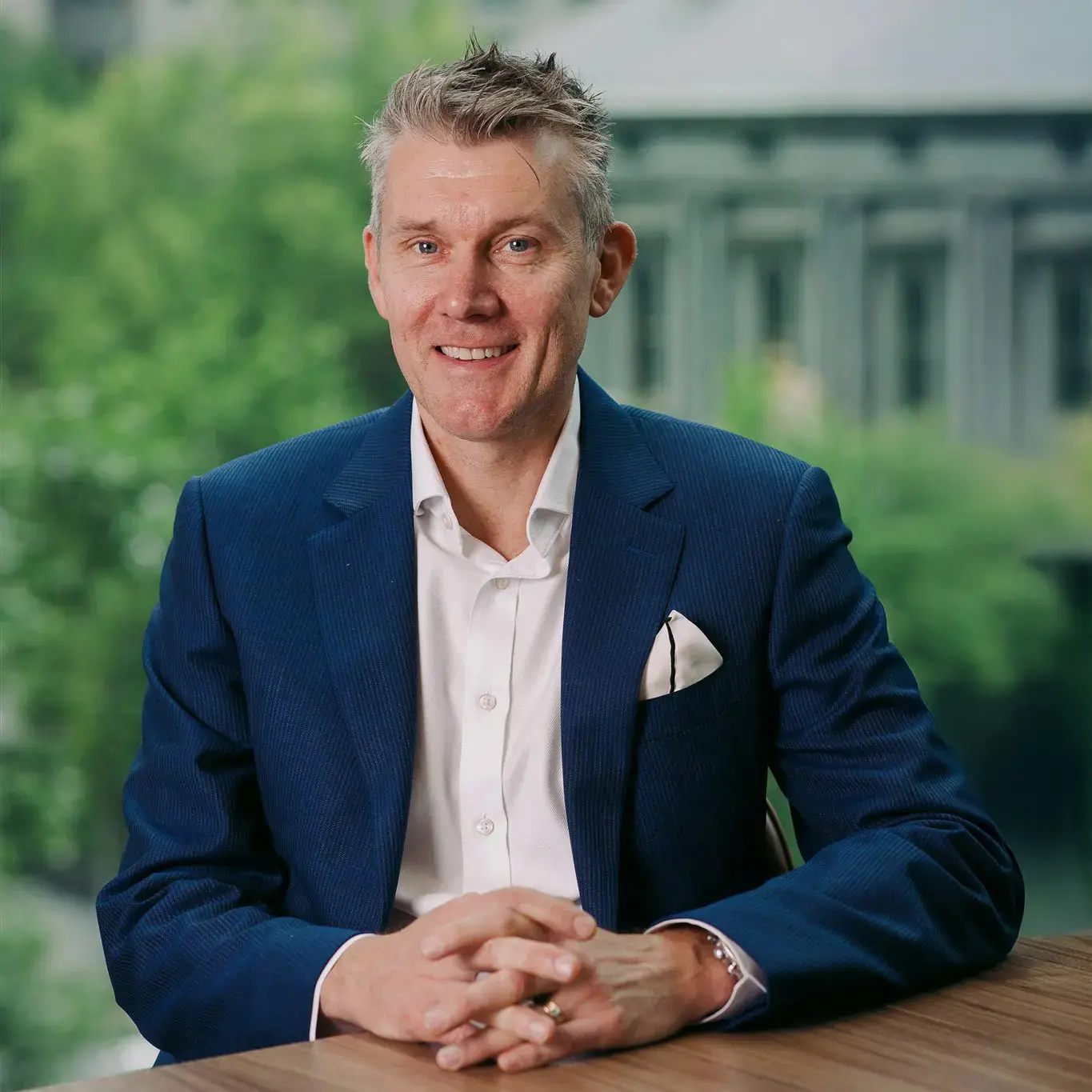 Professional portrait of smiling man seated at desk, with blurred trees and buildings in the background.