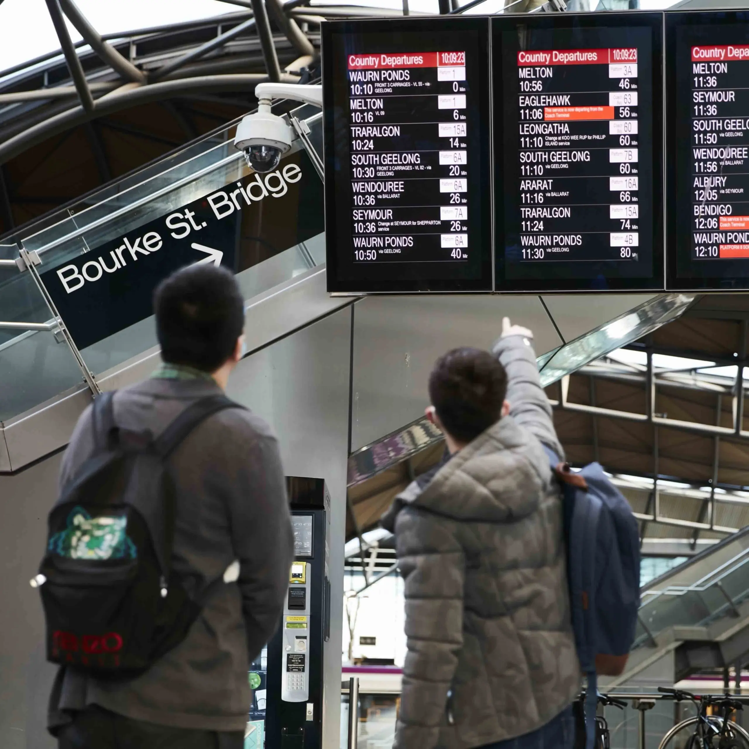 Two people, one pointing and the other observing, look at the digital train departure information board displaying train times for various stations. The Bourke St Bridge sign is visible in the background.