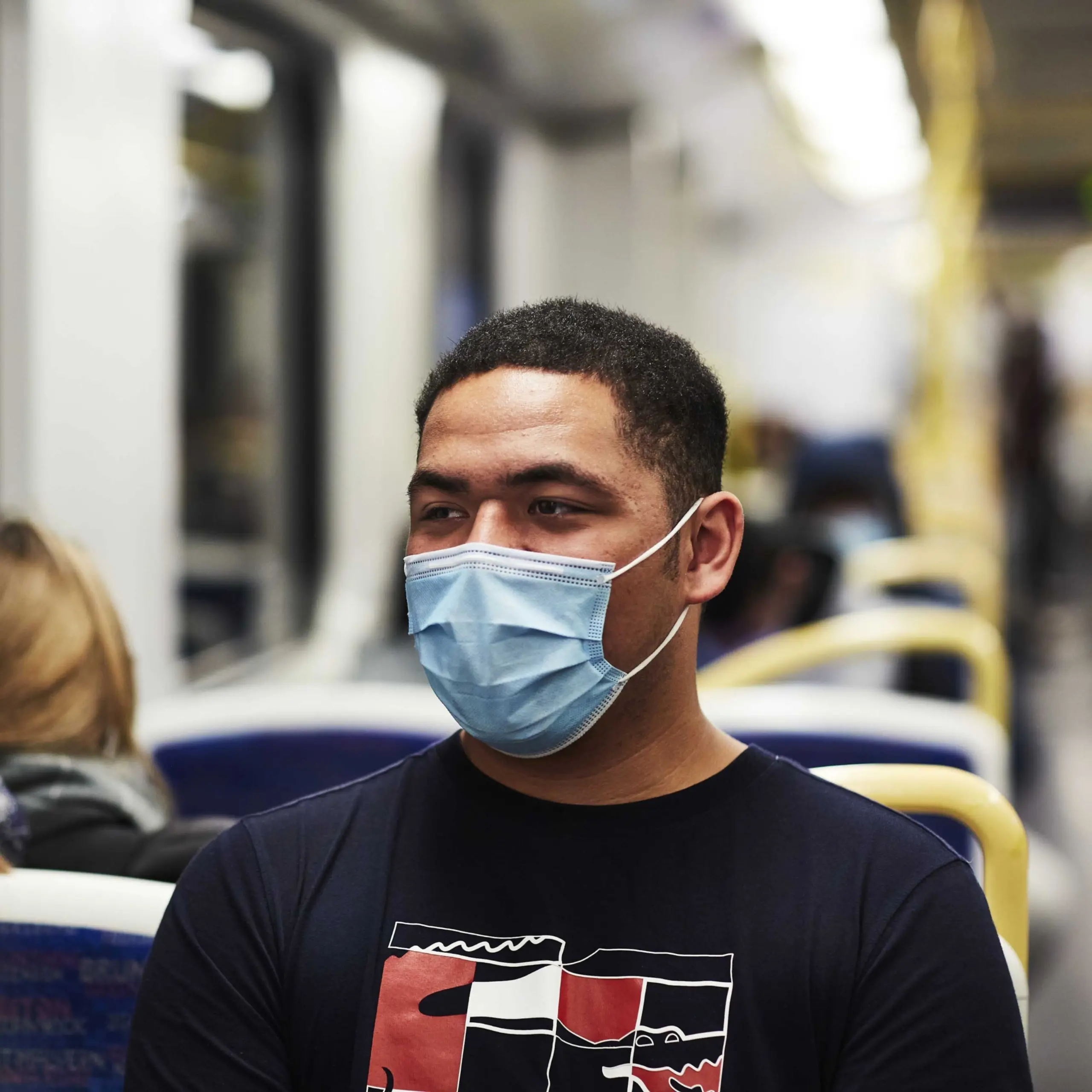 A close-up image of a passenger wearing a face mask and a navy blue t-shirt, sitting on a tram, looking ahead, with other passengers seated in the background.