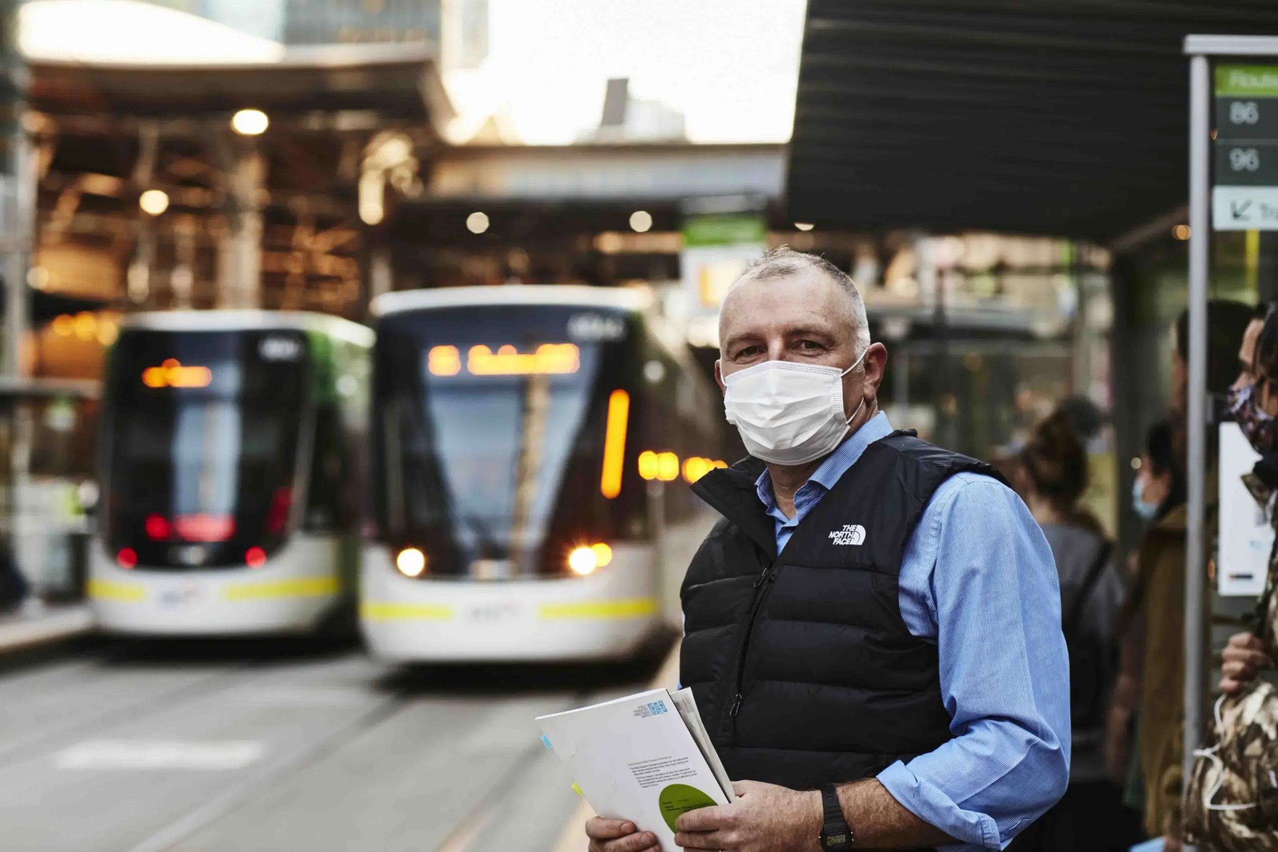 Simon McKenzie is standing at a tram stop, looking at the camera, wearing a mask and holding documents, with trams blurred in the background.