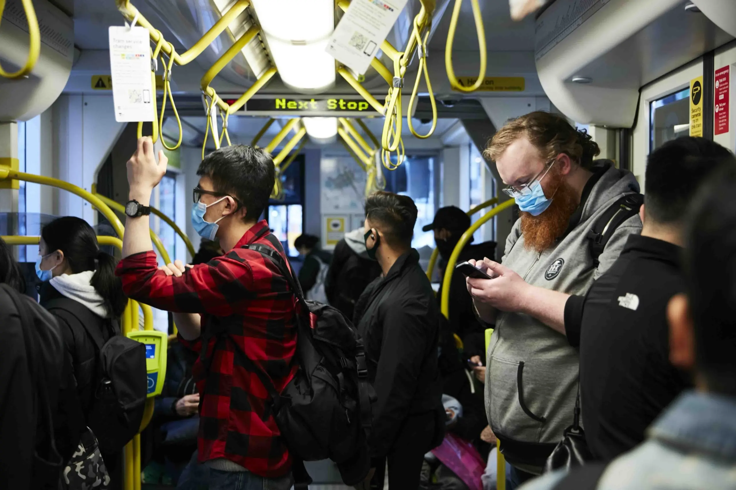 Several passengers stand inside a tram, all wearing masks. The image shows yellow tram handles and signs, with a 