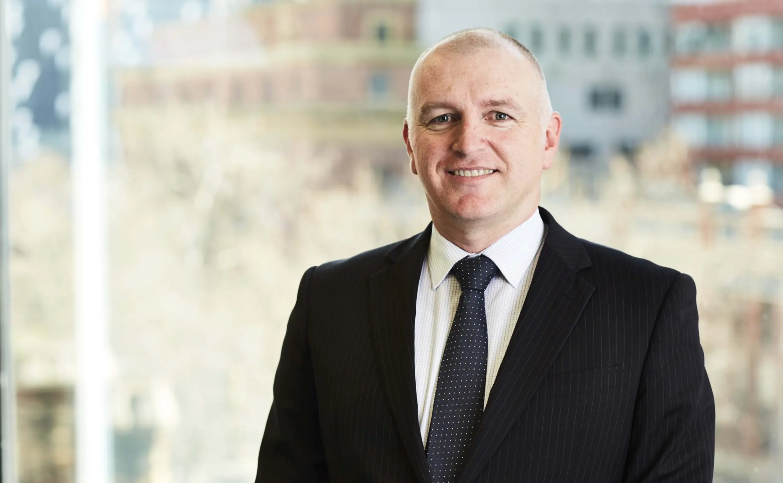 A professional portrait of Simon McKenzie, dressed in a black suit and tie, smiling at the camera, with a cityscape blurred in the background.