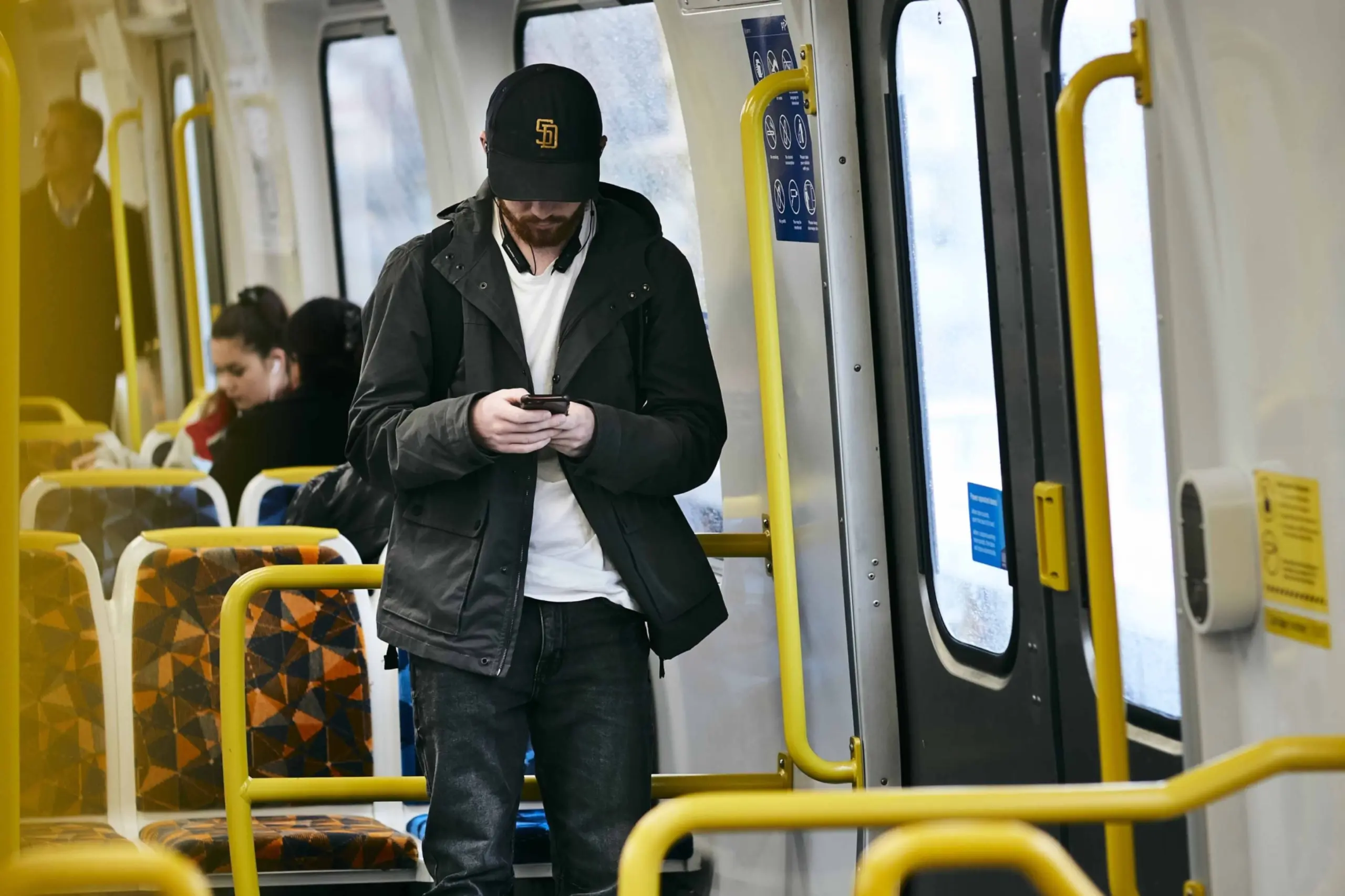 A man stands in a tram carriage, looking down at his phone, with other passengers seated in the background.