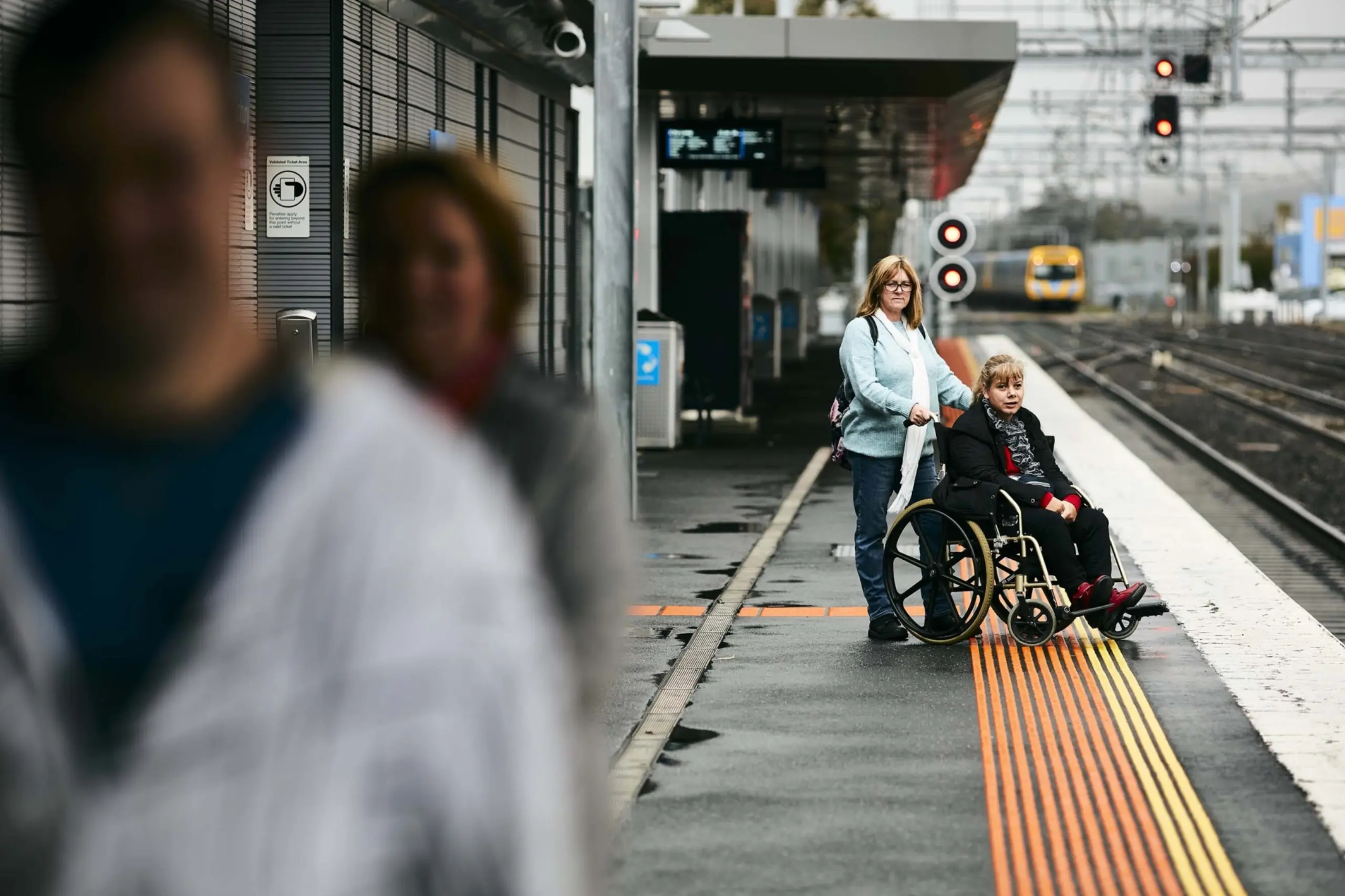 A woman stands behind another woman in a wheelchair at a train station. They are on the station platform with visible railway signals and a blurred train in the background.