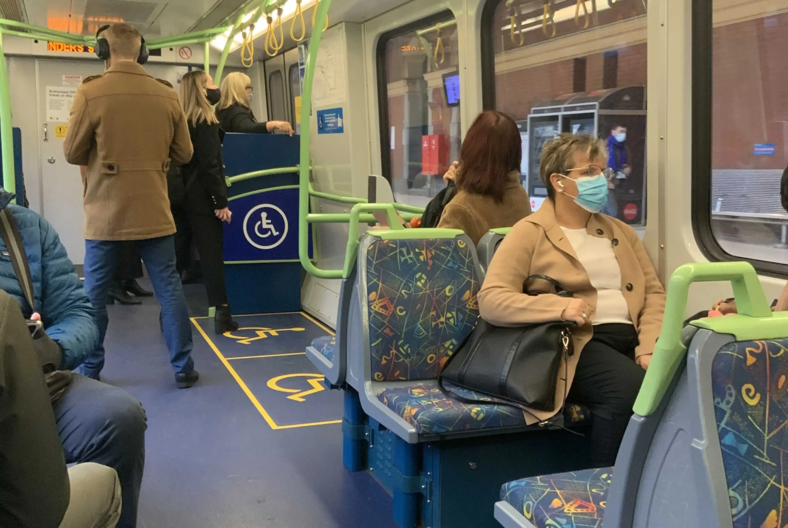 A middle-age woman wearing a mask and sitting on a train and looking out of the train window, with other seated and standing passengers behind in a clean, modern train interior.