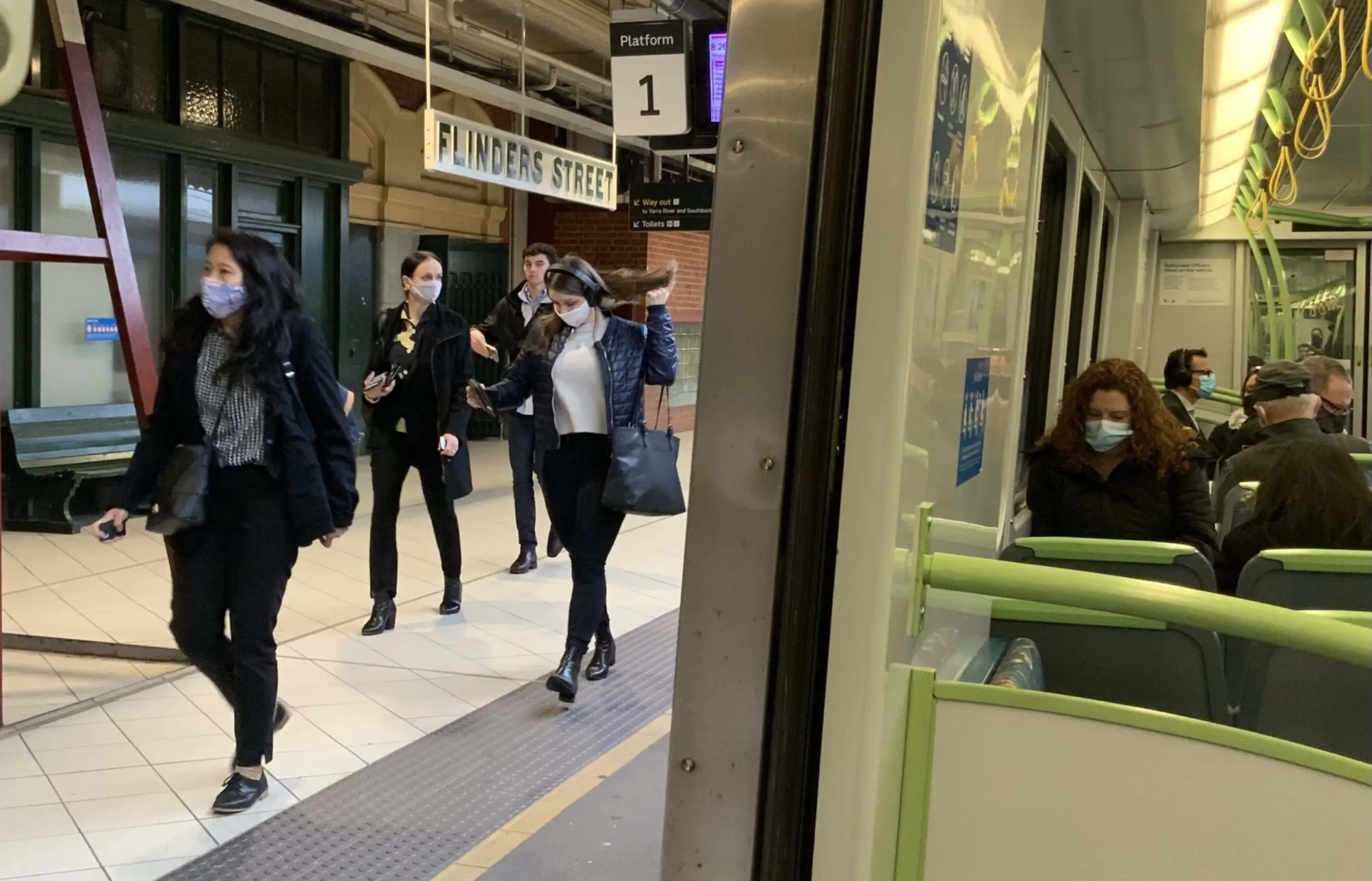 A train at Flinders Street Station with passengers wearing masks on the platform and inside the train, showing the station signage and urban background.