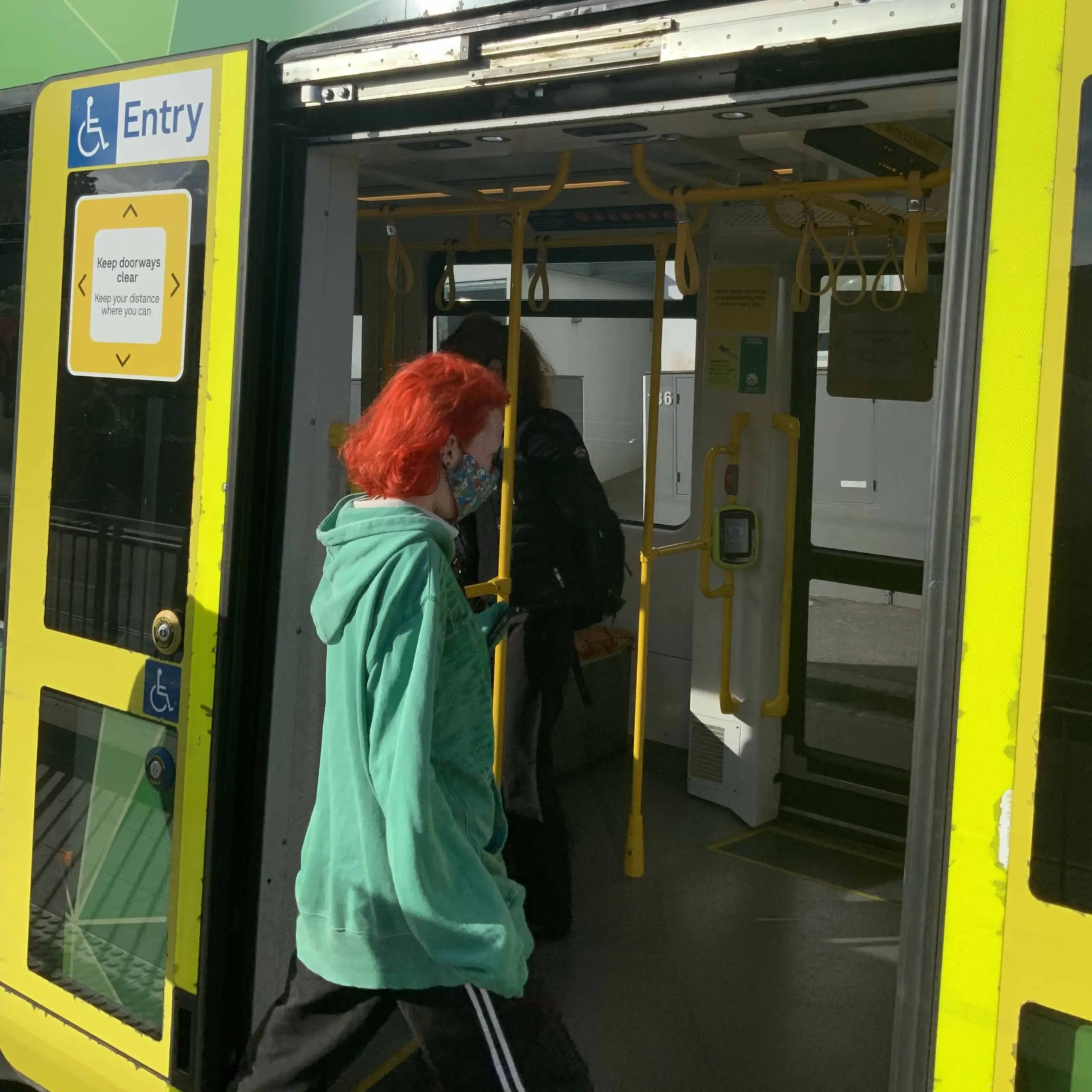 A passenger is walking towards the open door of a tram, with a wheelchair Entry sign on the door, and a brightly coloured green and yellow tram interior visible through the door.