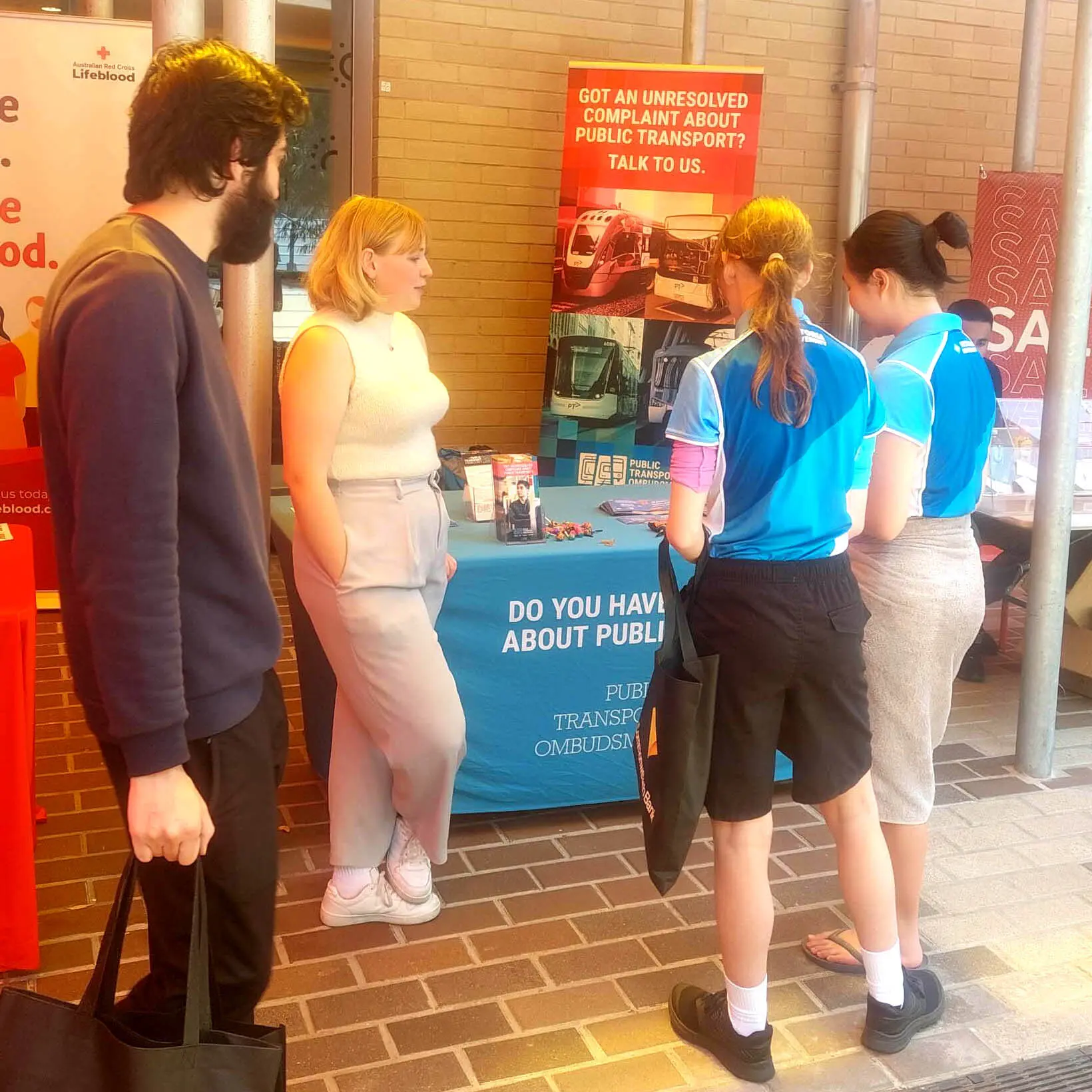 A group of students stands around a table at an event, where a Public Transport Ombudsman (PTO) staff member talks to them. The booth displays information about public transport complaints, with banners and pamphlets visible.
