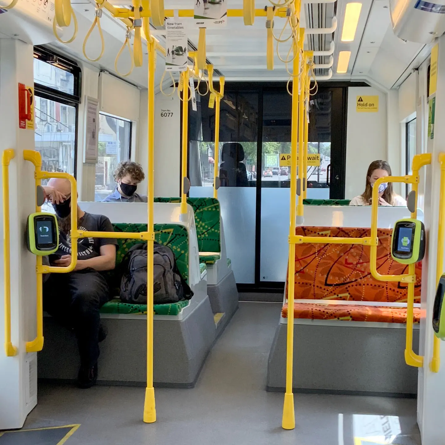 Inside a tram, passengers are separated and sitting in different seats in a tram carriage, following social distancing guidelines. The tram’s interior features yellow handrails and colourful seating.