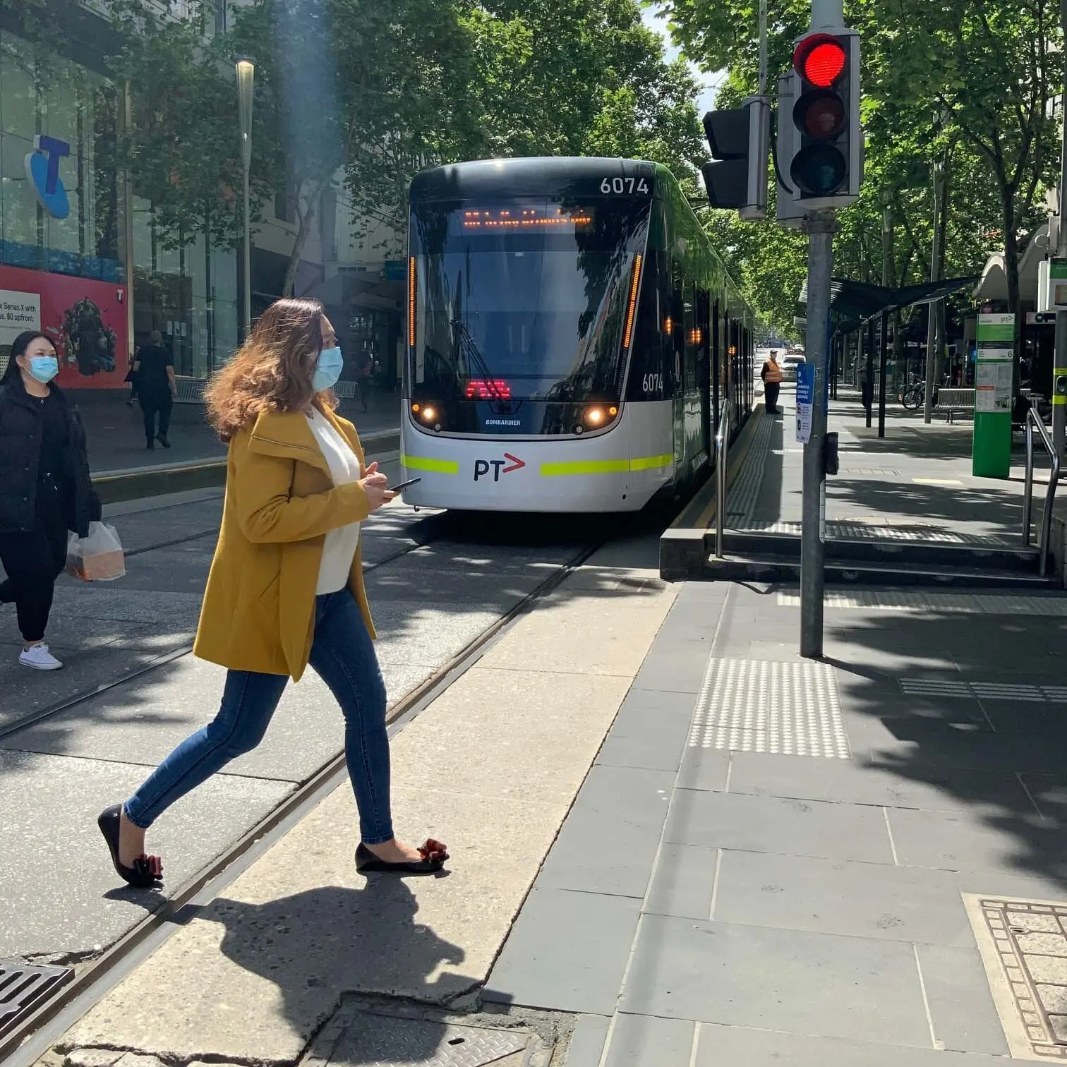 A pedestrian wearing a mask crosses a street near a tram stop, with buildings and public transport infrastructure and traffic light in the background.