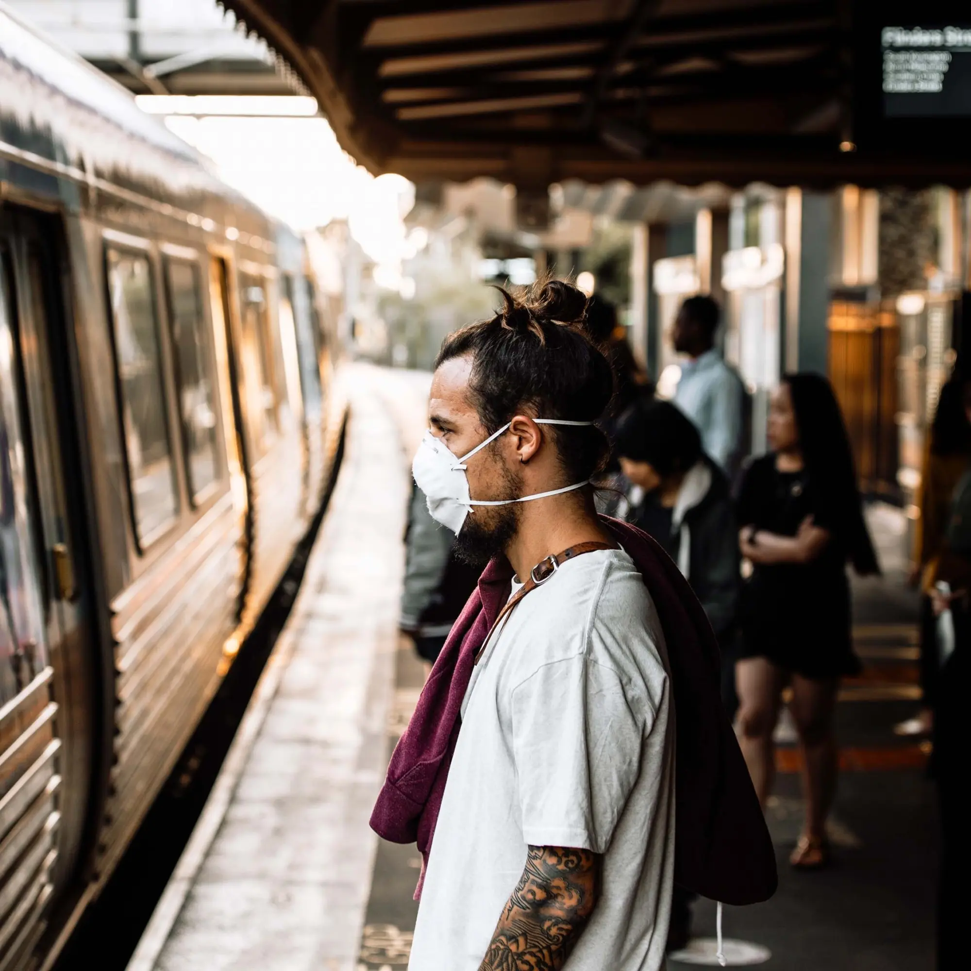 A close-up portrait of a man wearing a mask while standing at a train station platform and facing a train, with a blurred background of other people standing at the platform.