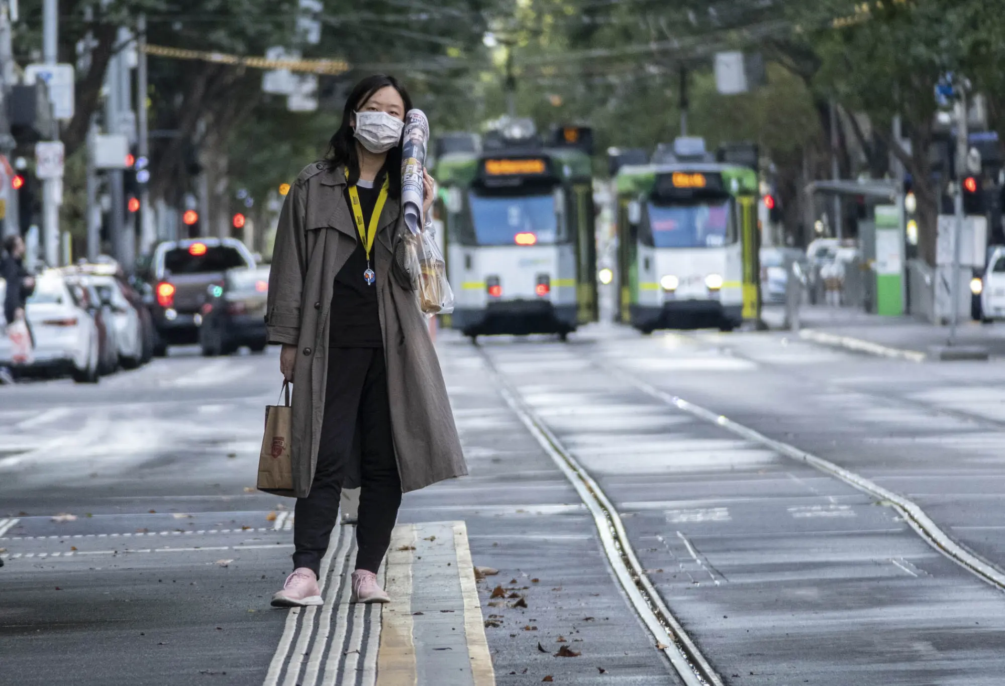 A woman, wearing a mask, stands next to the tram tracks holding a rolled-up newspaper, with trams arriving in the background. The image shows the bustling street scene with cars lining the street.