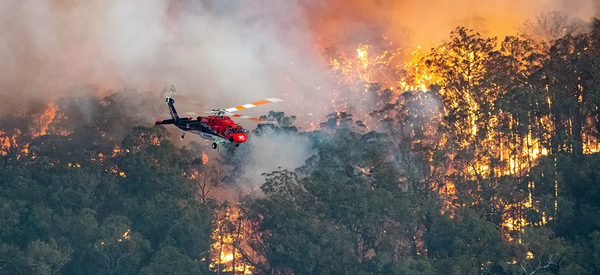 A CFA (Country Fire Authority) helicopter hovers above the forest as bushfires rage below in Mallacoota, Victoria. The landscape is engulfed in flames.