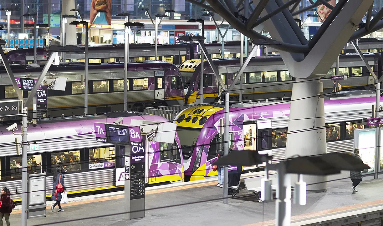 Image showing train station, with multiple purple and silver Metro trains sitting at the station with passengers visible inside and walking on the platforms. The station sign shows the train line number and destination.